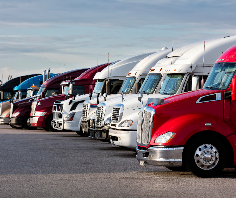 A row of semi-truck cabs in various colors, including red, maroon, and white, are parked closely together on asphalt