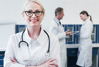 Smiling female doctor with glasses and stethoscope stands confidently in a clinic