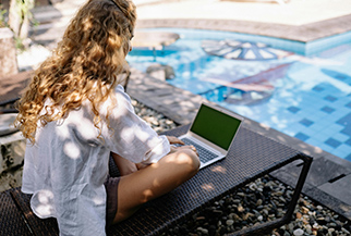 A woman with curly hair sits on a lounge chair by a pool, working on a laptop - www.mysoutherninsurance.com