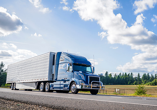 A blue semi-truck with a large trailer drives on a highway under a bright blue sky with scattered clouds - www.mysoutherninsurance.com