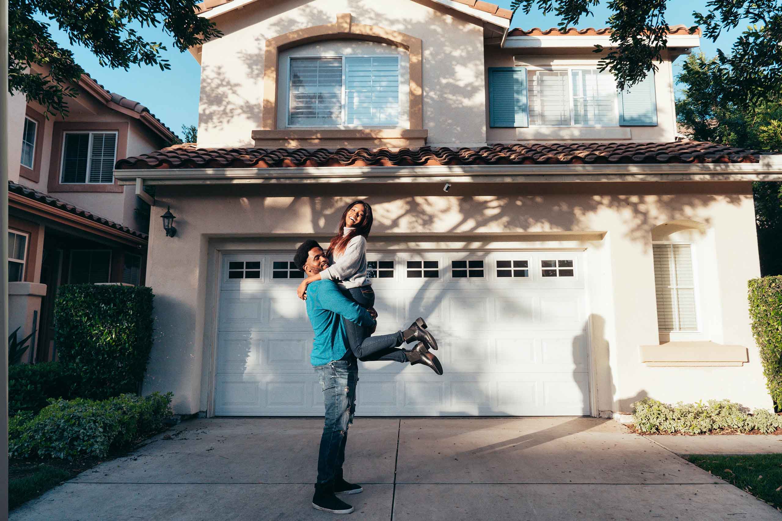A joyful couple stands on a driveway in front of a beige two-story house - www.mysoutherninsurance.com
