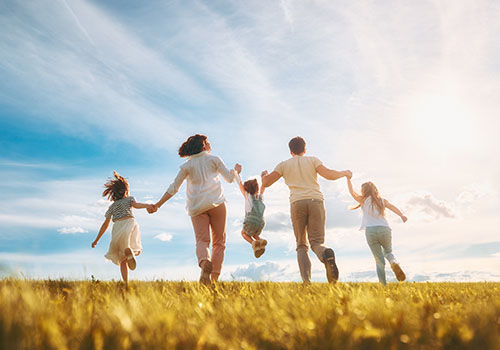 A family of five joyfully runs across a sunlit grassy field, holding hands - www.mysoutherninsurance.com