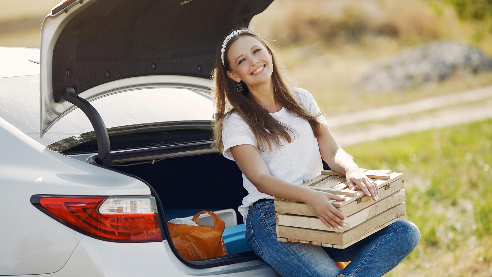 Woman sitting at the back of a car with open trunk, holding wooden crates outdoors on a sunny day - www.mysoutherninsurance.com