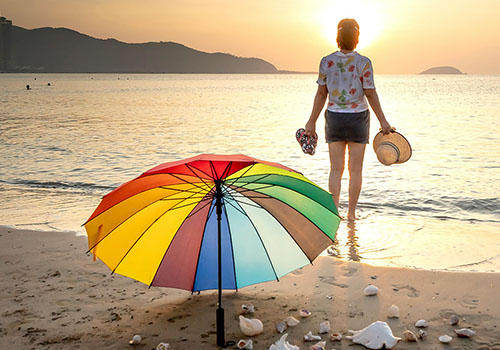 A person stands on a beach at sunset, holding sandals and a hat - www.mysoutherninsurance.com