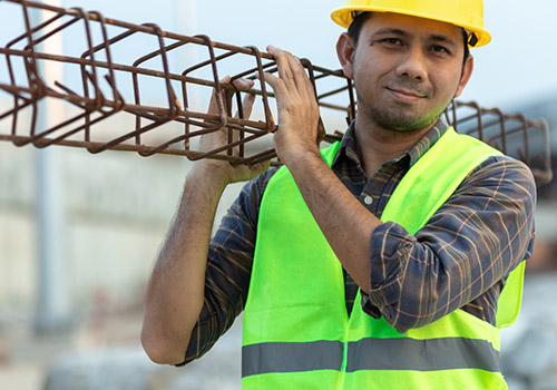 A construction worker wearing a yellow hard hat and neon safety vest carries a metal beam on his shoulder - www.mysoutherninsurance.com