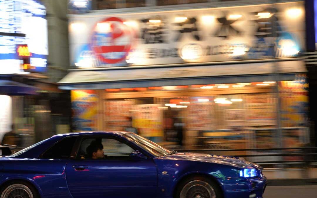 A blue car driving along a dimly lit street at night, with streetlights illuminating the surroundings